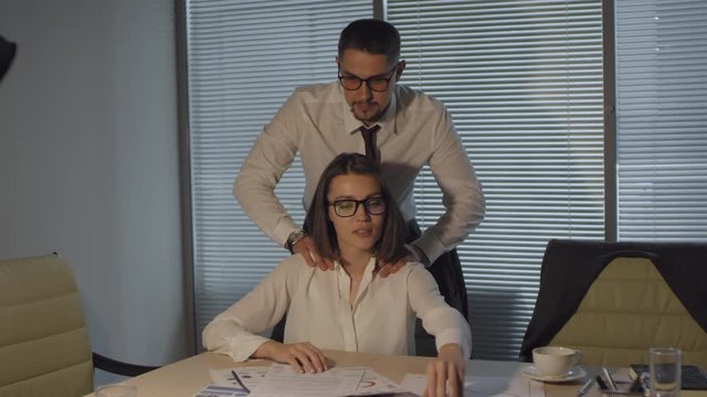 Medium shot of two young colleagues in business attire working together at night in dark office with closed blinds, smiling woman looking at charts and documents, and man giving her shoulder rub