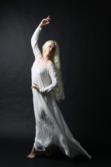 Ghostly full length portrait of a woman with long blonde hair wearing a white robe. Standing pose  against a black studio background. 