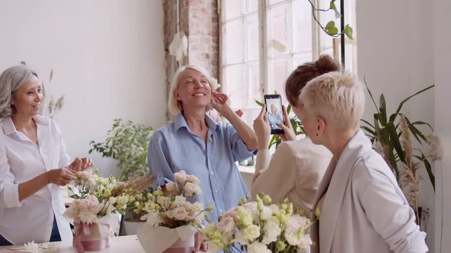 Medium shot of young female florist standing with smartphone in her hands and making photos of middle-aged blonde woman while two other women watching