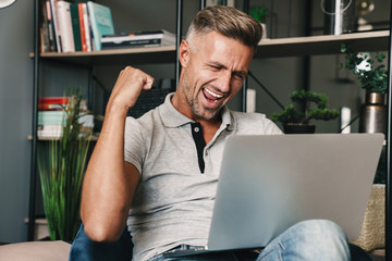 Photo of excited adult man smiling and celebrating while using laptop in apartment