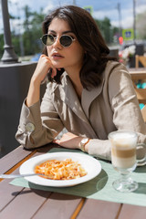 Beautiful smiling brunette girl wearing sunglasses is having lunch in a street cafe, sitting at a table and eating Italian pasta and drinking coffee. Advertising, commercial design. Copy space