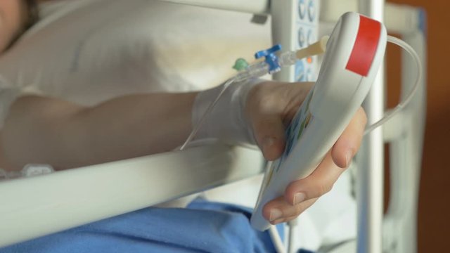 Patient with medical IV drip needle in the vein trying to press an emergency button and call for help on a hospital bed. A hospitalized young person with serious injuries feels pain and call a nurse.