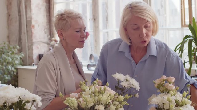 Tilt up of two blonde middle-aged women standing together at worktop in workshop, choosing flowers for flower composition and smelling them