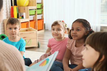 Kindergarten teacher reading book to cute little children indoors