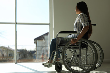 Young woman in wheelchair near window indoors. Space for text