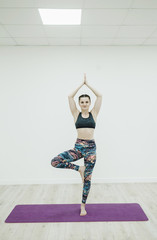 Attractive smiling woman doing yoga and standing in Tree pose on blue mat