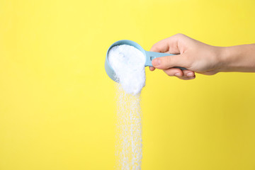 Woman pouring laundry powder from measuring scoop on yellow background, closeup