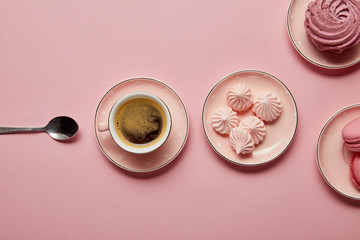 Top view of pink meringues and macaroons on pink dotted saucers with spoon and cup of coffee on pink background