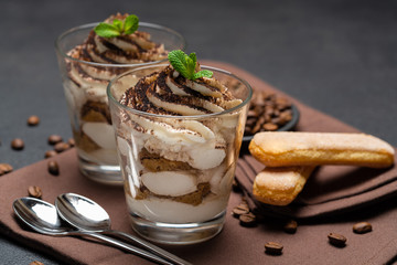 Classic tiramisu dessert in a glass cup on brown kitchen towel or napkin on dark concrete background