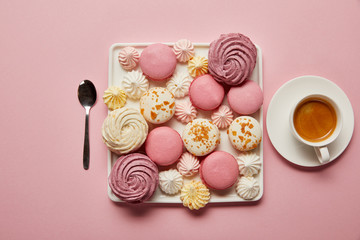 Fototapeta na wymiar Flat lay with assorted meringues and macaroons on square dish with cup of coffee and spoon on pink background