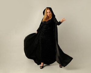 full length portrait of blonde woman wearing long black flowing clock and lace gown, standing pose against a creamy studio background.