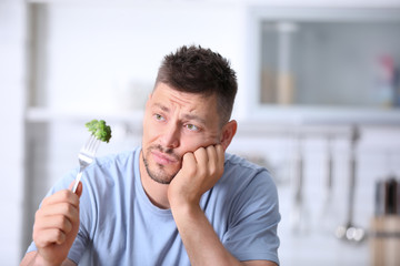 Portrait of unhappy man looking at broccoli on fork in kitchen - obrazy, fototapety, plakaty
