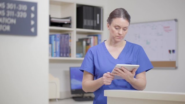 Nurse, framed right, reviews the patient record on a tablet at station.