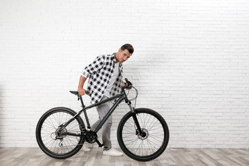 Handsome young man with modern bicycle near white brick wall indoors