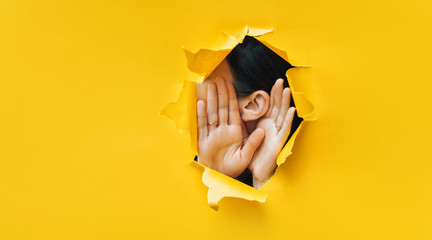 Female ear and hands close-up. Copy space. Torn paper, yellow background. The concept of eavesdropping, espionage, gossip and the yellow press. - obrazy, fototapety, plakaty