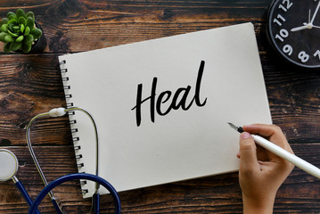 Top view of stethoscope,plant, clock and hand holding pen writing Heal on notebook on wooden...