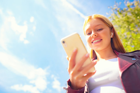 Young beautiful smiling adult Caucasian girl walking outdoor in the park having chat communication with wireless headphones mobile reading talking listening using with her hand on sunset at summer