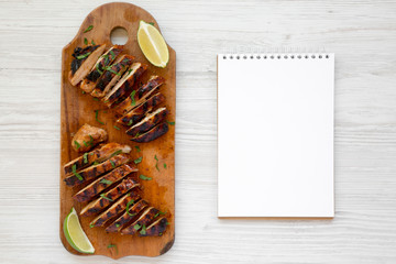 Grilled chipotle chicken breast on a rustic wooden board, blank notepad on a white wooden surface, top view. Flat lay, overhead, from above. Copy space.