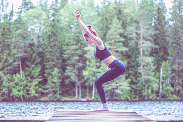 Young yogi  girl  practicing yoga, standing in Utkatasana exercise, Chair pose on the lake.  Concept of healthy life and natural balance