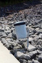 The metal garbage can in the rock pile on a sunny day.
