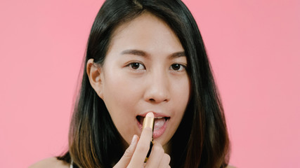 Naklejka premium Young beautiful fashionable Asian woman applying lips makeup with cosmetic brush in casual clothing over pink background studio shot. Happy smiling adorable glad woman rejoices success.