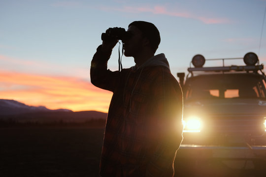 A man with binoculars near the car at sunset. Man Binoculars Looking Mountain Cloudscape Traveling Concept