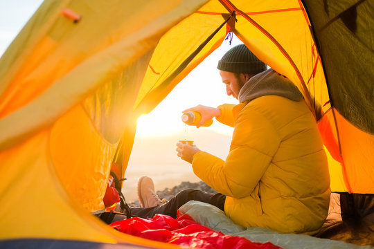 the traveler guy in a yellow jacket and in a tent, woke up and pours himself tea from a thermos