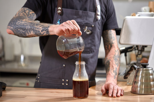 Man""s hand with tattoo pour freshly made coffee into glass bottle on a wooden table.