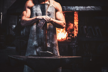Muscular blacksmith in protective apron at his workshop with hammer in his hands. There are fire at background.