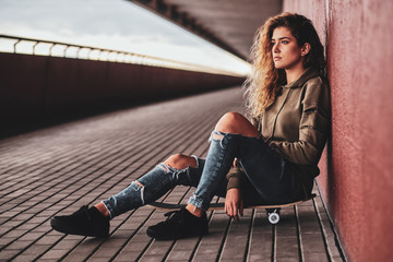 Beautiful young woman in ripped jeans is sitting on her own skateboard at the street.