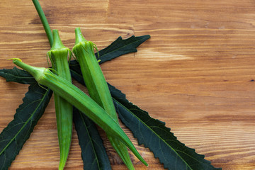 Three green ripe okras lie on a sheet of okra in dark green color on the left on a brown background, on the right is an empty place, top view