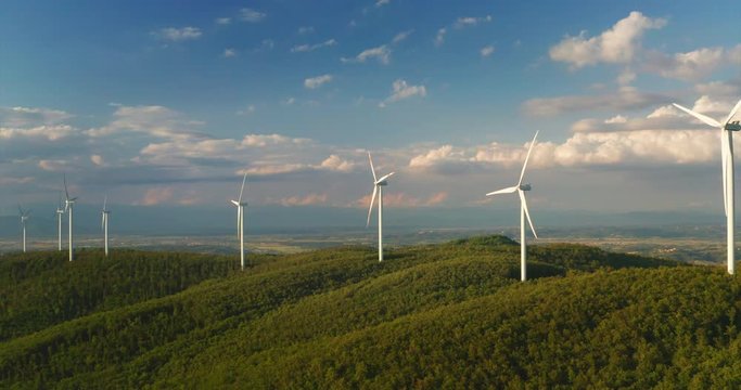 Aerial shot of wind mills rotating by the force of the wind and generating renewable energy in a green ecologic way to the planet with a sun shining.