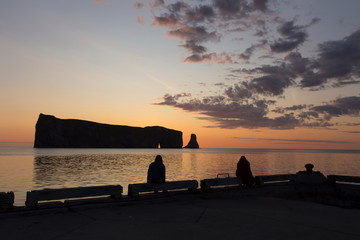 Two women seen in silhouette sitting on the Percé pier watching the sun rising behind the famous rock during a beautiful summer morning, Quebec, Canada