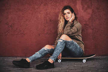 Beautiful young woman in ripped jeans is sitting on her own skateboard at the street.
