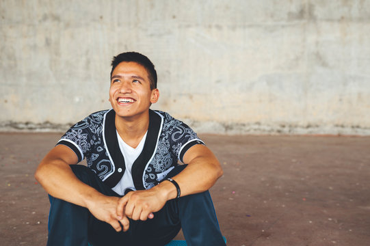Portrait of hispanic young break dancer seated in urban park with concrete wall on background