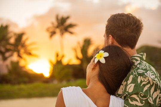 Romantic couple relaxing watching sunset on beach stroll view from back. Woman resting head on lover's shoulder on honeymoon vacation travel in summer Hawaii destination. Newlyweds people.