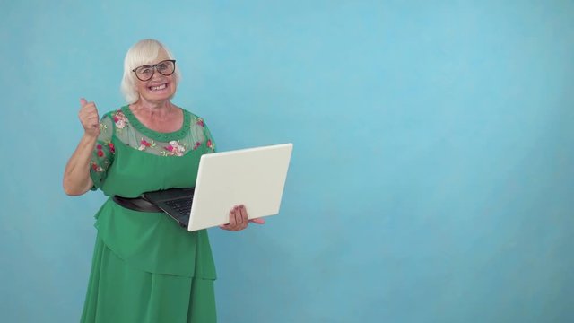 Joyful positive elderly woman with a laptop in her hands learned about the win on a blue background copy space