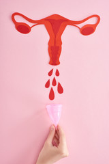 cropped view of woman holding menstrual cup near red paper cut female reproductive internal organs with blood drops on pink background