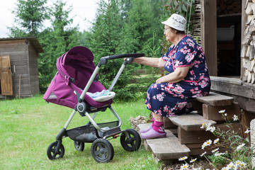 Senior woman sitting on timber house porch with baby carriage, caring great-grandchild on backyard