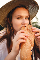 Portrait of beautiful brown haired girl in straw hat eating baguette bread while dreamily looking in camera on picnic in city park