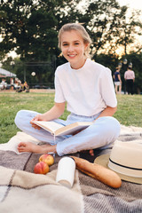 Young beautiful smiling woman sitting on plaid with book happily looking in camera on picnic in park