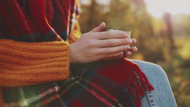 Close up of woman`s hands holding cup of hot tea. Tourist girl wearing tartan scarf enjoys warm drink in autumn park