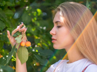 slender charming young blonde in white T-shirt and colorful sundress poses in front of camera against background of fruit tree in city park. Sunny day in big city. Chinese apples in woman's hand