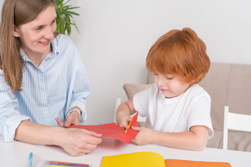 Obraz na płótnie Canvas Little red-haired boy with a nanny or mother or teacher cuts out of colored paper with scissors.