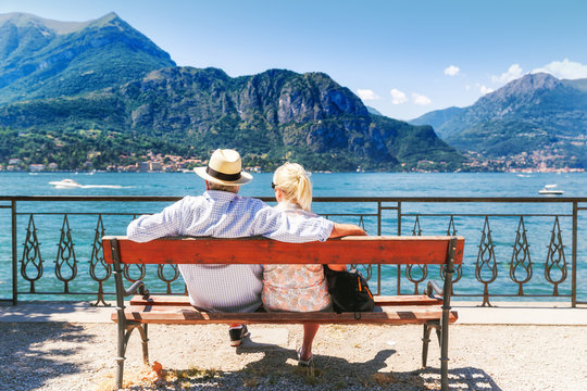 Lake Como, village Bellagio, Italy. Senior couple weekend getaway having rest on the bench by spectacular lake Como in Italy. Sunny day scenery. Tourists admiring view on popular tourist attraction.