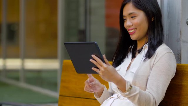 leisure, technology and people concept - smiling asian woman with tablet computer sitting on bench in city yard