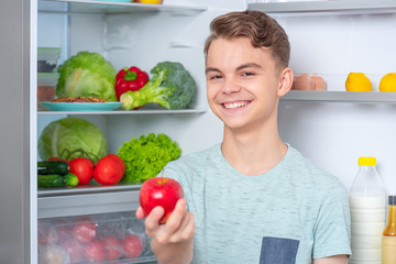 Smiling handsome young teen boy holding fresh red apple while standing near open fridge in kitchen at home. Portrait of pretty child choosing food in refrigerator full of healthy products.