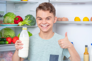 Smiling handsome young teen boy holding bottle of milk and drinks over eyes while standing near open fridge in kitchen at home. Portrait of child choosing food in refrigerator full of healthy products