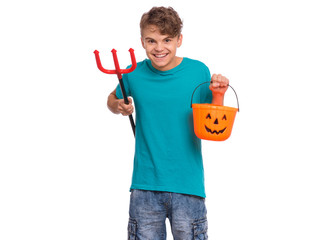 Happy halloween concept. Smiling teen boy holding orange pumpkin bucket and red trident, isolated on white background. Joyful young teenager looking at camera. Funny child waiting candy.