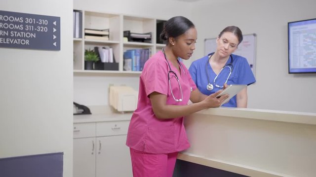 Two nurses w/ stethoscopes at nurses station review chart, camera move right